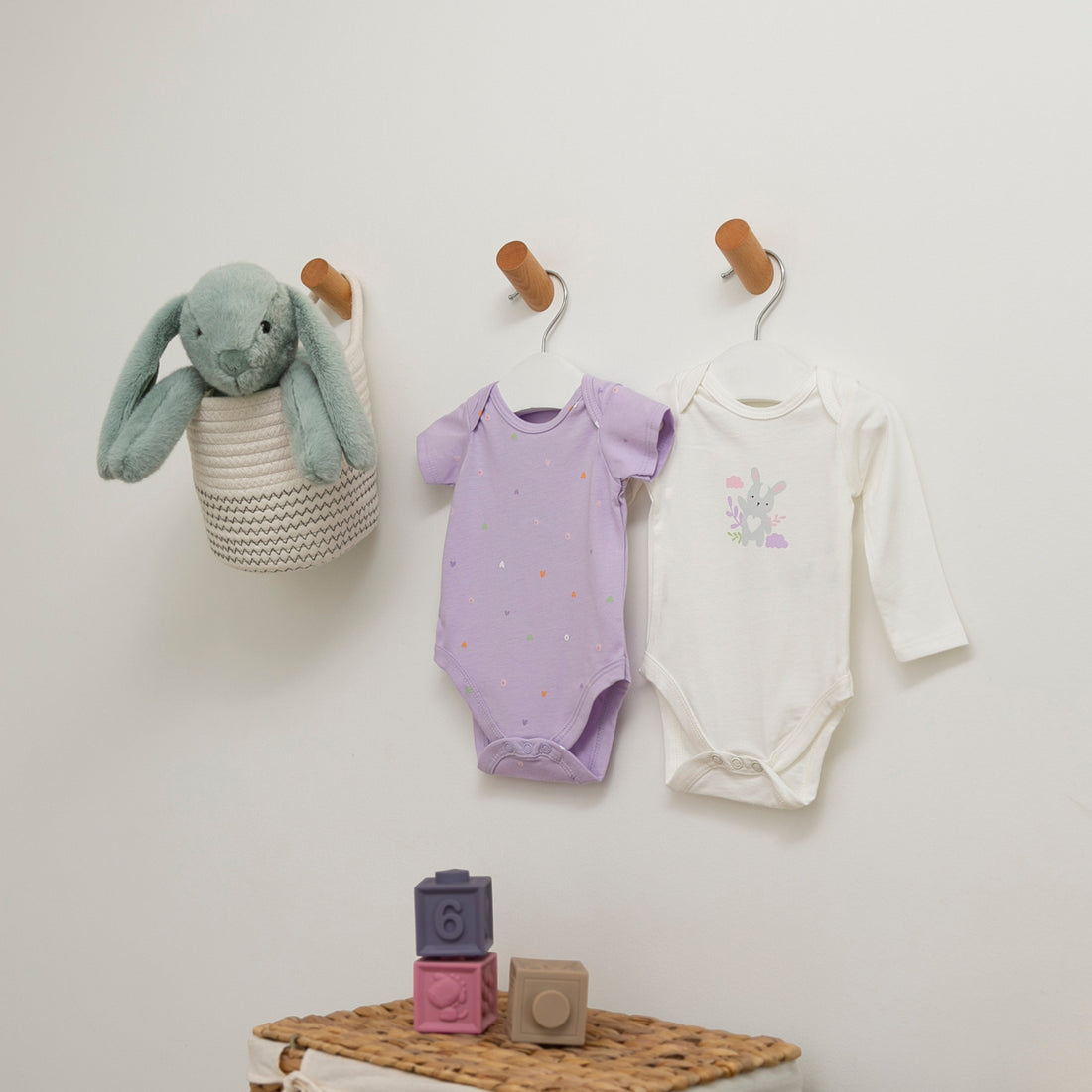 Zwei Bodys an einer Kinderkleiderstange, neben einem Kleiderkorb und einigen Babyspielzeugen.