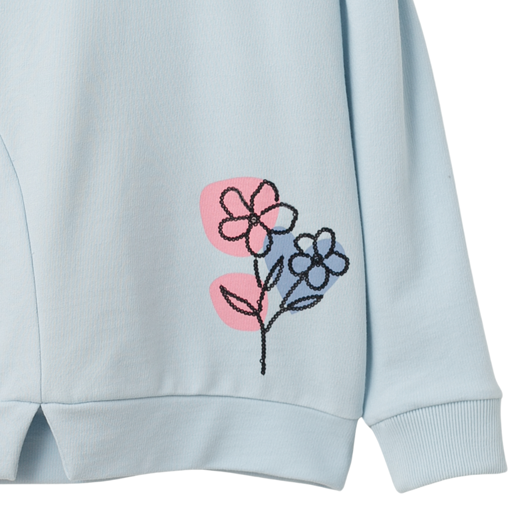 Nahaufnahme der zarten Paillettenblume und des feinen Bundschnitts des hellblauen Mädchenpullovers.